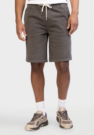 Hombre lleva pantalones cortos verde oliva con cordón, camiseta blanca, calcetines blancos y zapatillas beige, de pie frente a un fondo liso gris claro.