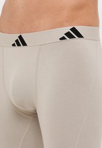 Calções de desporto bege com uma textura suave e um design ajustado. Apresenta uma cintura com riscas pretas e o logótipo da marca na parte da frente.
