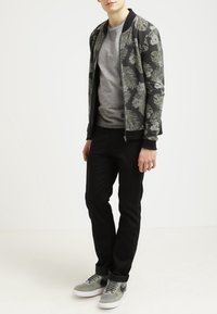 Giovane in giacca bomber nera floreale, t-shirt grigia, pantaloni neri e sneaker grigie che sta in piedi di fronte a uno sfondo bianco semplice.