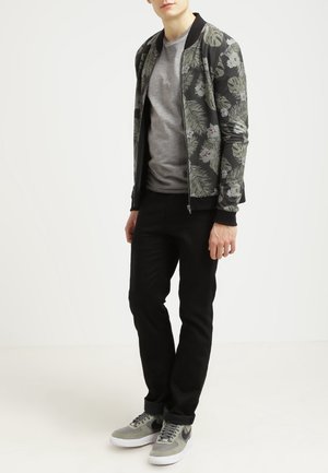 Giovane in giacca bomber nera floreale, t-shirt grigia, pantaloni neri e sneaker grigie che sta in piedi di fronte a uno sfondo bianco semplice.