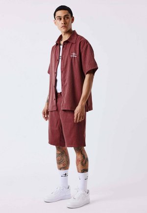 Junger Mann mit kurzen Haaren, der ein bordeauxfarbenes Kurzarmshirt und Shorts trägt, weiße Sneakers und sichtbare Bein-Tattoos, steht vor einem schlichten Hintergrund.