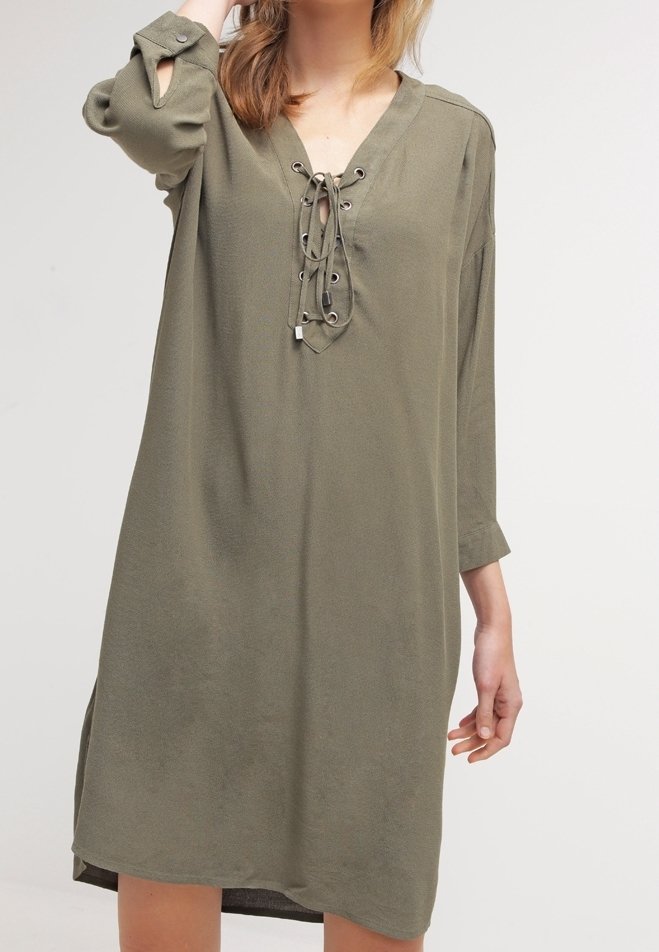Femme portant une robe ample mi-longue couleur olive avec un décolleté en V lacé et des manches trois-quarts, sur un fond uni.