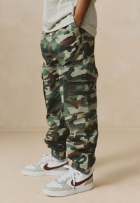Camouflage-Cargohosen in Grün und Braun mit mehreren Taschen. Elastische Bündchen und lockere Passform, kombiniert mit weißen und braunen Nike-Sneakers.