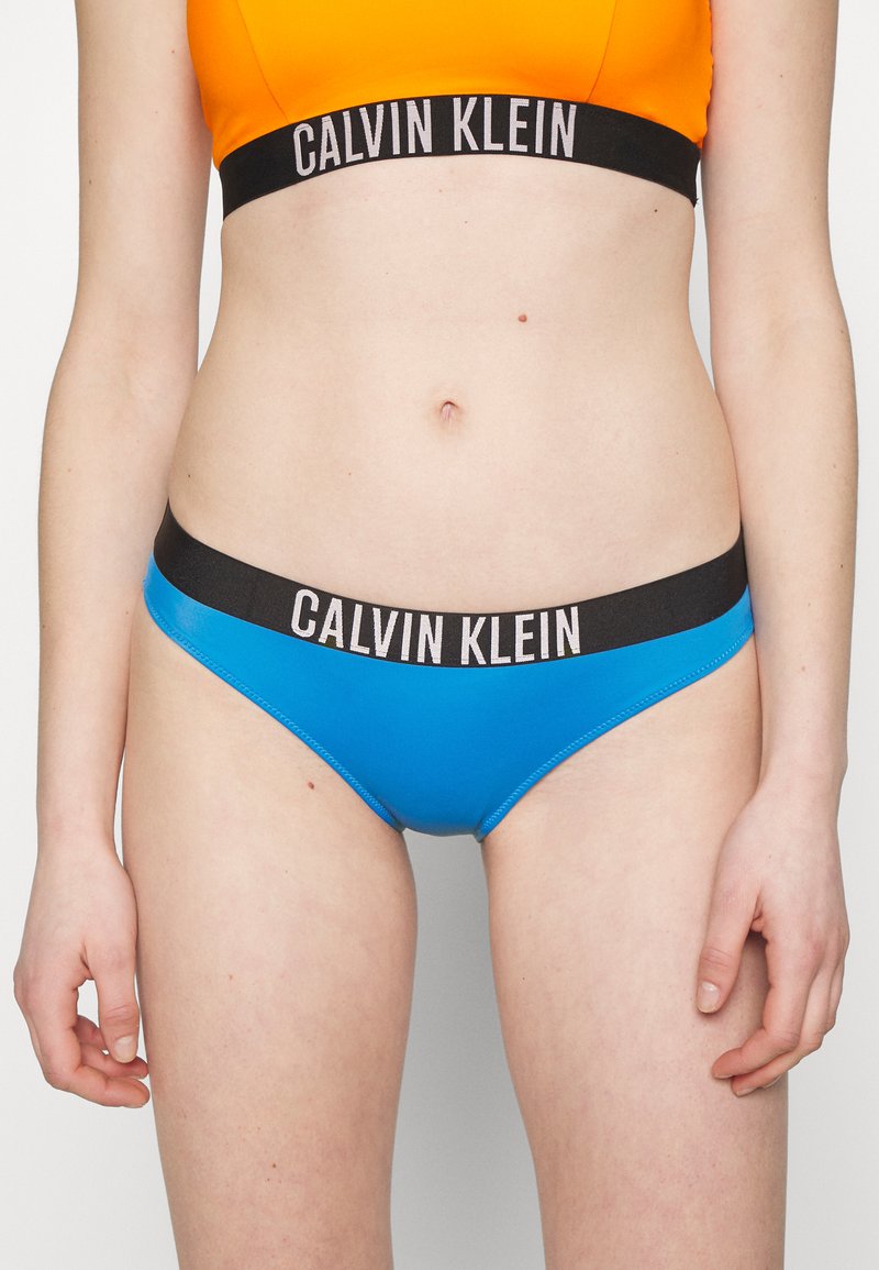 Bikini blu con fascia nera e branding "CALVIN KLEIN" bianco. Texture liscia, taglio a vita bassa e design minimalista.