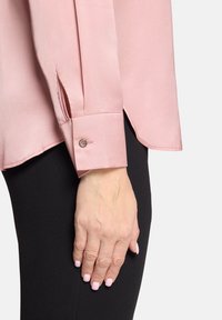 Jasnoróżowa satynowa bluzka z długimi rękawami, mankietami na guziki, bocznymi rozcięciami i gładką fakturą; noszona na czarnym materiale.