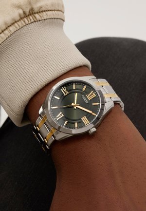 Silver and gold-toned watch with a green dial, Roman numerals, and a stainless-steel band, worn on a wrist with a beige sleeve.