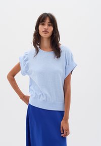 Femeie cu părul închis la culoare, de lungime medie, purtând un top scurt cu mânecă scurtă de culoare bleu deschis și o fustă albastră, pe un fundal alb simplu.