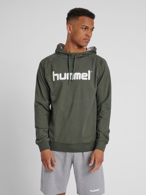 Ung mand i olivengrøn hættetrøje med "hummel" logo og grå shorts, der kigger til siden mod en ensartet lys baggrund.