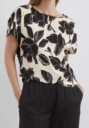 Femme portant un haut beige à manches courtes avec un grand imprimé floral abstrait noir et un pantalon noir, les mains dans les poches.