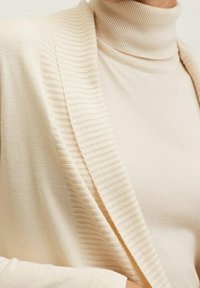 Cardigan beige côtelé porté sur un pull à col roulé beige côtelé assorti, visible de l'épaule jusqu'à mi-torse.