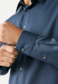 Mężczyzna zapinający guzik mankietu na ciemnoniebieskiej, teksturowanej koszuli z długim rękawem i kołnierzykiem, zbliżenie na dłonie i szczegóły koszuli.