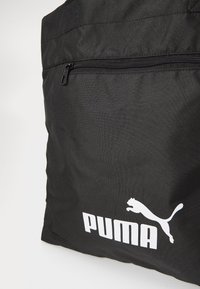 Melnas auduma somiņa ar rāvējslēdzēja kabatu un izteiksmīgu baltu Puma logotipu ar lecošu panteru un treknu tekstu.