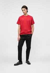 Młody mężczyzna stoi w czerwonej koszuli z krótkim rękawem, czarnych spodniach i czarnych mokasynach na jednolitym jasnoszarym tle.