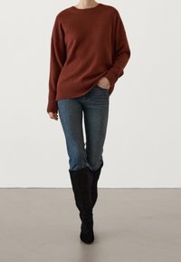 Un pull en tricot couleur rouille avec une coupe décontractée, associé à un jean bleu ajusté et des bottes noires jusqu'aux genoux. Texture lisse et ourlet côtelé.