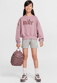 Sudadera morada de Nike con logotipo naranja, pantalones cortos grises, calcetines rosas y blancos, y mochila marrón con patrones blancos. Zapatillas deportivas grises con detalles rosas.