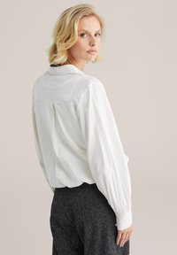 Camicia bianca con bottoni, dal taglio morbido, caratterizzata da maniche lunghe con polsini arricciati e un sottile design a yoke sul retro.