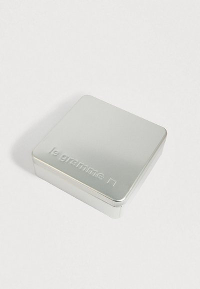 Srebrna kovinska škatla s zaobljenimi koti, ki ima na pokrovčku embosirano blagovno znamko "le gramme", nameščena na beli površini.