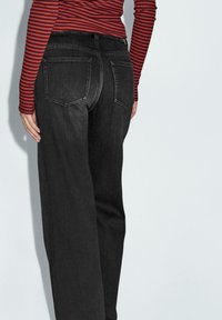 Jeans in denim nero con taglio dritto, dotati di due tasche posteriori e vita media, abbinati a una maglietta a maniche lunghe a righe rosse e nere.