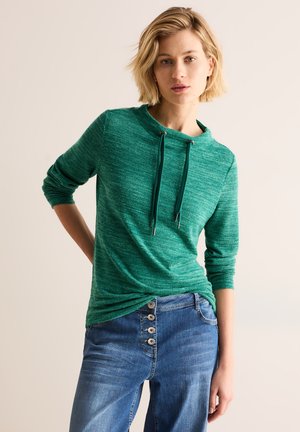 Sweatshirt vert, texturé, avec capuche et cordon de serrage. Manches longues, coupe décontractée, associé à un jean en denim bleu avec des accents de boutons.