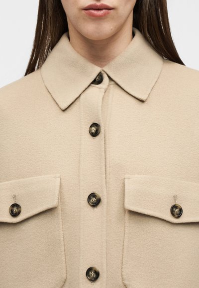 Γυναίκα που φοράει ένα μπεζ πουκάμισο-σακάκι από μαλλί με μυτερό γιακά, μεγάλα σκούρα κουμπιά και δύο μπροστινές τσέπες με καπάκι και κουμπιά.