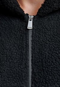 Primo piano di una cerniera argentata su un tessuto di pile nero testurizzato, con il tirante della cerniera inciso con la parola "Dropsize".