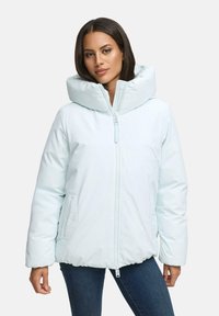 Donna che indossa un giubbotto imbottito azzurro chiaro con colletto alto e zip frontale, abbinato a jeans blu scuro, in piedi contro uno sfondo semplice.
