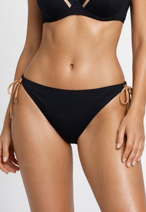Kobieta w czarnym bikini z beżowymi wiązaniami po bokach, ukazująca środkową część ciała i dłonie na jednolitym jasnym tle.