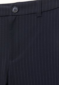 Gros plan sur un pantalon de costume bleu marine à fines rayures, avec un bouton noir, une boucle de ceinture et un détail de poche avant.