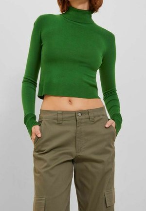 Woman wearing a fitted green turtleneck cropped sweater and olive cargo pants, with hands in pockets, standing against a plain background.