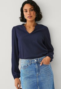 Top lungo blu navy con maniche a sbuffo, finitura testurizzata e increspata; presenta un collo a V discreto ed è abbinato a una gonna di denim.