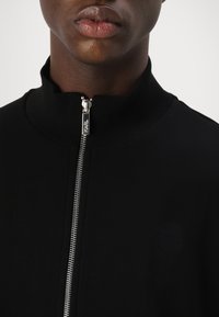 Schwarze Zip-Jacke mit hohem Kragen, ausgestattet mit einem silbernen Reißverschluss und einem dezenten, geprägten Logodesign. Glattes, leichtes Material.