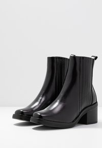 Botines negros brillantes con tacones de bloque, lados elásticos acanalados y tiradores, exhibidos sobre una superficie blanca con un fondo gris liso.