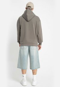 Mężczyzna stojący tyłem, ubrany w taupe'owy hoodie, beżową czapkę, jasnoniebieskie długie dżinsowe krótkie spodenki, białe skarpetki i białe trampki na białym tle.