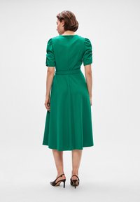 Πράσινο φόρεμα μέχρι το γόνατο με φουσκωτά μανίκια, στενή μέση και φερμουάρ. Λείο ύφασμα με μονόχρωμο σχέδιο και χωρίς ορατά σχέδια.