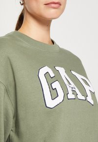 Felpa verde oliva con un grande logo "GAP" ricamato in bianco e blu navy. Il tessuto è morbido, con scollo a girocollo e una vestibilità comoda.