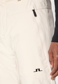 Beige cargo bukser laget av glatt stoff, med en glidelåslomme og en svart logo aksent på benet.