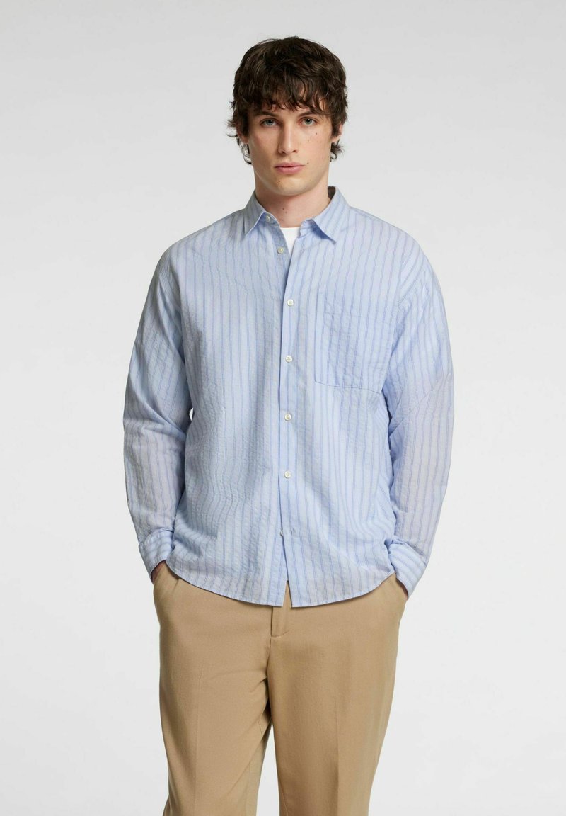 Camicia a maniche lunghe azzurro chiaro con righe verticali, colletto a bottoni e taschino a sinistra, abbinata a pantaloni beige.