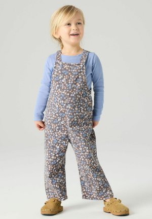 Copil mic zâmbitor cu păr blond, purtând o bluză albastră cu mâneci lungi, salopetă maro cu flori și pantofi slip-on bej, stând pe un fundal simplu.