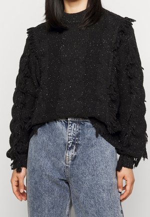 Vrouw draagt een zwarte, grofgebreide trui met franjes aan de mouwen en blauwe, hooggelede spijkerbroek.