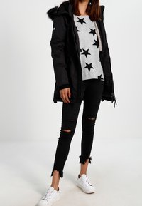 Zwarte parka met nep-bont capuchon, grijze sweater met sterrenmotief, versleten zwarte jeans en witte sneakers. Kenmerken zijn onder andere ritsen en trekkoorden.