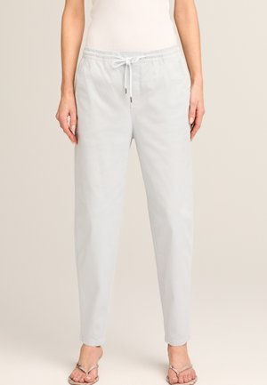 Frau trägt hellgraue, mit Kordelzug versehene Hose und offene Sandalen, steht vor einem einfarbigen beigen Hintergrund.