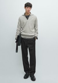 Hellgrauer Strickpullover mit Reißverschlusskragen, kombiniert mit schwarzer Hose. Hält eine schwarze Ledertasche. Glatte Textur und lockere Passform.