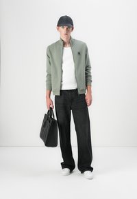 Light green zip jacket over a white shirt, paired with dark wide-leg jeans. Accessories include a black cap and black bag, with white sneakers.