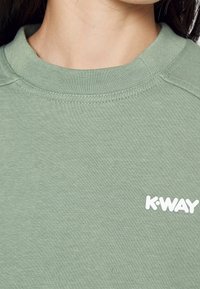 Une personne portant un sweatshirt K-WAY vert clair, montrant le décolleté et le logo sur la poitrine.