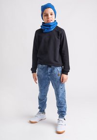 Chłopiec stoi w niebieskiej czapce i szaliku, czarnej bluzie, niebieskich dżinsach i białych trampkach na jednolitym białym tle.