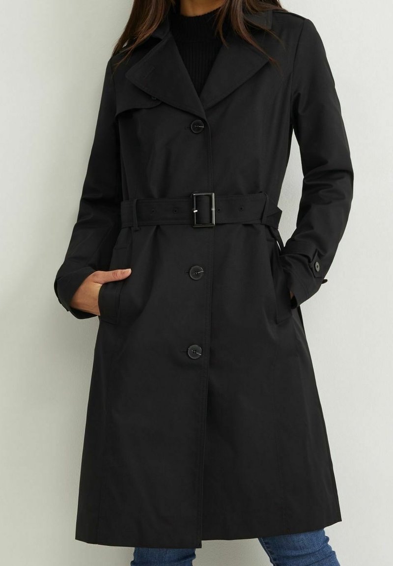 Femme portant un long trench-coat noir ceinturé avec des boutons, debout les mains dans les poches, portant un jean bleu en dessous.