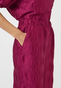 Violet samettihousut, joissa on teksturoitu aaltoileva kuvio, joustava vyötärö ja sivutaskut. Lähikuva korostaa materiaalia ja suunnittelua.