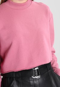 Mujer vistiendo un suéter corto rosa combinado con pantalones de cuero negro de cintura alta y un cinturón negro con una hebilla plateada.