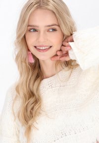 Cremefarbener gestrickter Pullover mit strukturiertem Muster, lockerer Passform und weit geschnittenen Ärmeln. Lange, wellige blonde Haare und pinkfarbene Quastenohrringe.