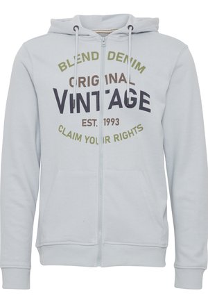 Jasnoniebieski rozpinany welurowy polar z miękkiego materiału, z frontową kieszenią, sznurkami oraz nadrukowanym tekstem w ciemnym brązie i zieleni.